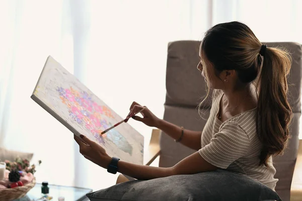 Jeune femme détendue assise sur un fauteuil à la maison et peignant sur toile avec des peintures à l'huile. — Photo