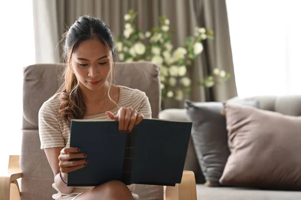 Pacífico asiático mujer leyendo libro mientras sentado en beige sillón en su apartamento. — Foto de Stock