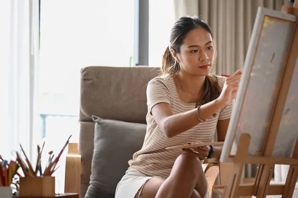 Jovem mulher casual sentada na frente do cavalete e pintura de quadro em tela. — Fotografia de Stock