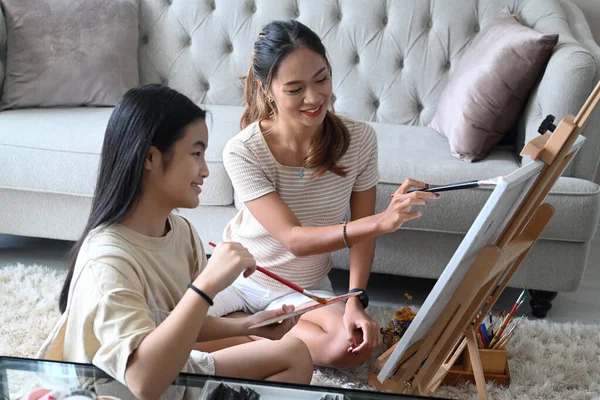 Smiling asian woman and her young sister sitting in front of easel and painting picture on canvas. — Stock Photo, Image