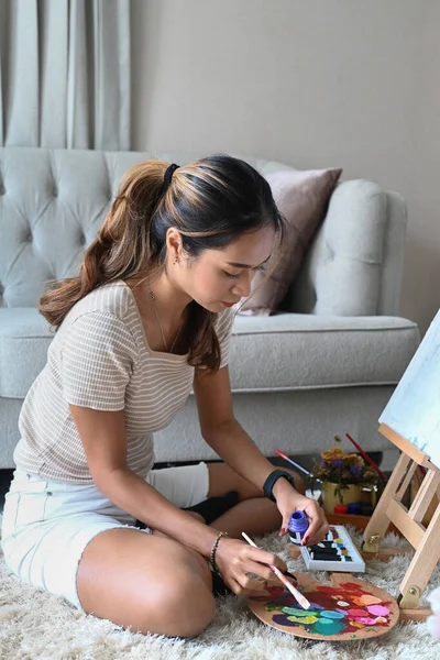 Creative young woman sitting in front of easel and painting picture with water color. — Stock Photo, Image