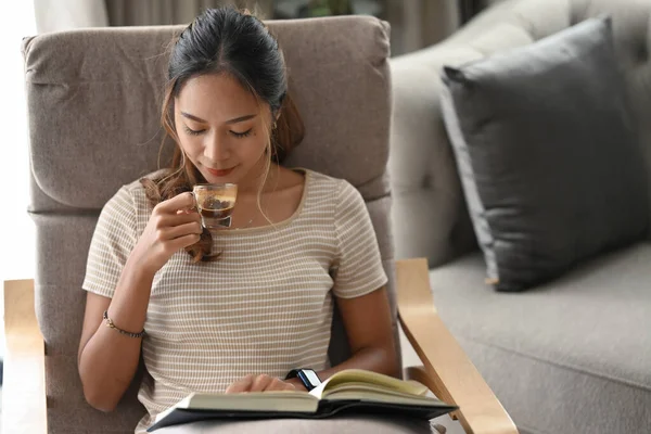 Satisfecho asiático mujer bebiendo café y leyendo libro en sillón en casa. — Foto de Stock
