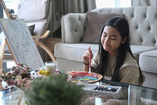 Jolie jeune fille asiatique en vêtements décontractés assis dans le salon et peinture tableau avec aquarelle. — Photo