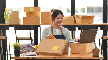 Gülümseyen küçük işletme sahibi kadın müşterilere teslimat için karton kutuda ürün paketlerken dizüstü bilgisayar kullanıyor. Online satış, e-ticaret