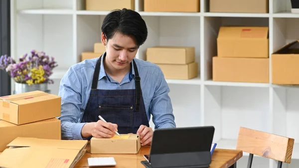 Young man online seller writing address on cardboard box while prepare parcel box of product for deliver. Concept of online selling, e-commerce