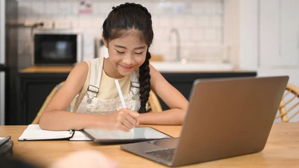 Симпатичная азиатская девушка дистанционно обучается онлайн, выполняя домашнюю работу, сидя за кухонным столом — стоковое фото