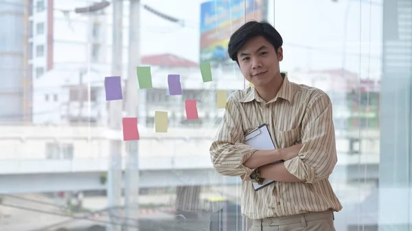 Giovane uomo d'affari fiducioso in piedi vicino a una grande finestra in ufficio con edifici della città in background. — Foto Stock