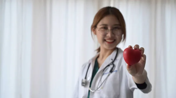 Glimlachende vrouwelijke arts in wit medisch uniform met rode hartvorm in de hand. Cardiologie, gezondheidszorg en medisch concept. — Stockfoto