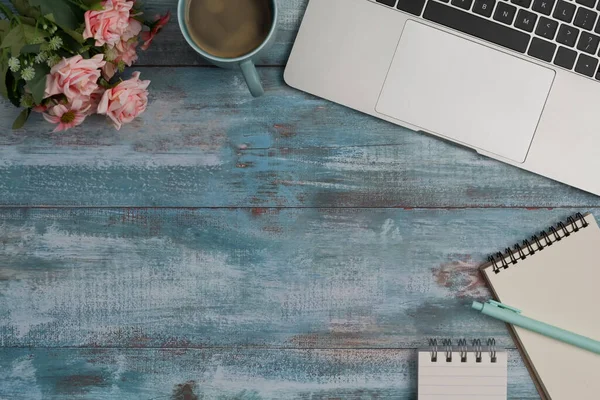 Elegante lugar de trabajo con computadora portátil, bloc de notas, taza de café y flores de color rosa en el escritorio de madera azul. —  Fotos de Stock