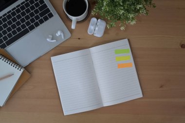 Üst görünüm açık defter, kablosuz kulaklık, dizüstü bilgisayar ve kahve fincanı ahşap masada..