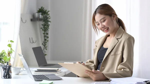 オンラインプロジェクトで働く女性従業員の笑顔、彼女のオフィスデスクで財務報告書を書く. — ストック写真