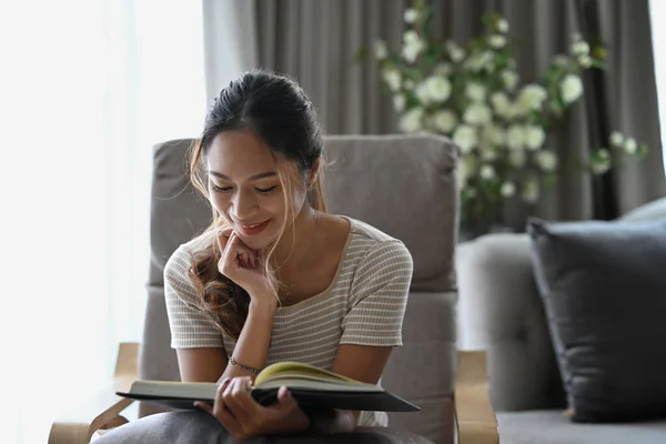 Felice donna millenaria lettura libro mentre seduto sulla poltrona beige in appartamento. — Foto Stock