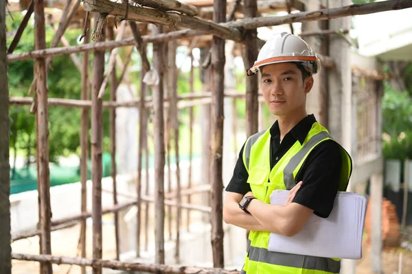 Attractive male inspector, supervisor or construction architect in helmet and safety reflective vest standing with crossed arms at construction site.
