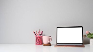 Bilgisayar dizüstü bilgisayarı, kahve fincanı, kalem tutacağı ve beyaz masada saksısı olan kadınsı bir iş yeri..