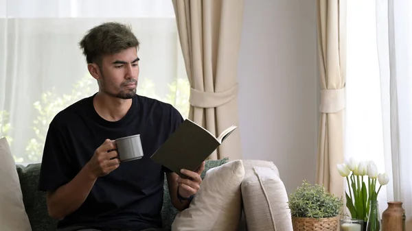 熱いコーヒーを飲むハンサムな男と家でソファに本を読んでください — ストック写真