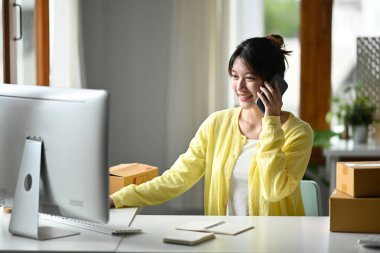 Gülümseyen genç bir kadın cep telefonuyla konuşuyor ve bilgisayardan ürün satın alma siparişini kontrol ediyor. E-ticaret, çevrimiçi satış kavramı. 