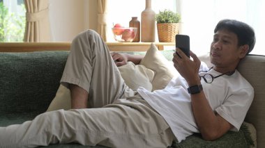 Evdeki kanepede uzanmış rahat Asyalı adam akıllı telefondan sosyal medyayı kontrol ediyor..