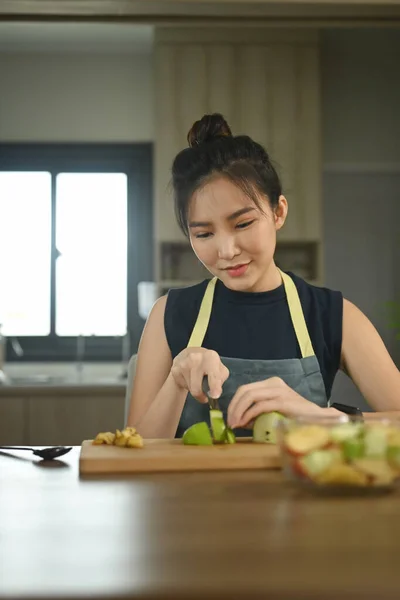 Happy woman cutting fruits on wooden board while making healthy salad breakfast in kitchen.