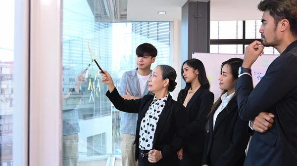 Focused senior female team leader and diverse businesspeople brainstorming together with sticky notes on glass window in a modern office.