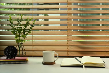 Kahve fincanı, ev bitkisi, kitaplar ve beyaz masa üzerindeki çalar saatli şık bir iş yeri..