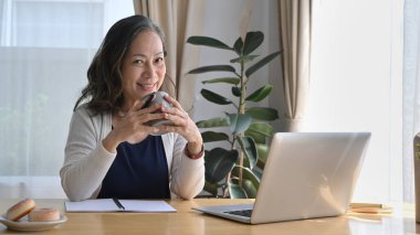 Çekici, olgun bir kadın elinde bir fincan çay tutarken uzaktan kumandayla kameraya gülümsüyor..