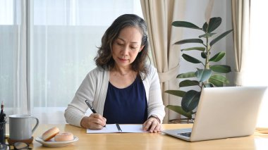 Odaklanmış olgun bir kadın dizüstü bilgisayarda internetten web sitesi izliyor ve evden uzakta çalışırken deftere not alıyor..