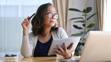 Gözlüklü düşünceli olgun bir kadın dijital tablet tutuyor ve evden çalışırken pencereden bakıyor..