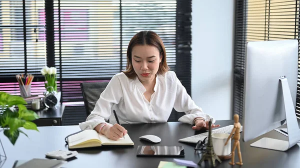 コンピュータの前に座ってノートにビジネスプランを書く集中アジアの女性ビジネス起業家 — ストック写真
