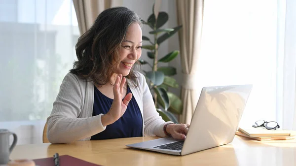 Smiling mature woman waving hand during video conference while remote working from home.