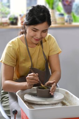 Önlüklü gülümseyen genç kadın atölyede el yapımı çömlekçi yaratmaktan zevk alıyor. Evin içinde yaşam tarzı aktivitesi, el işi, hobi konsepti..