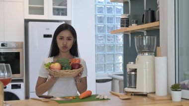 Neşeli Asyalı kadın elinde sepetle taze organik sebzeler ve meyvelerle modern ev mutfağında duruyor..