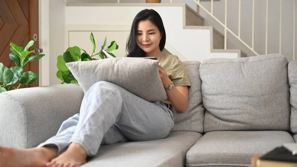 Relaxed young woman in casual clothes using digital tablet on couch, spending leisure time at home.