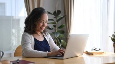 Orta yaşlı bir kadın rahat bir evde masada oturuyor ve gülen yüzlü bir bilgisayar kullanıyor..