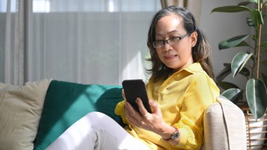 Kanepede dinlenen orta yaşlı, gülümseyen bir bayan sosyal ağdaki arkadaşlarına cep telefonundan mesaj yazıyor..