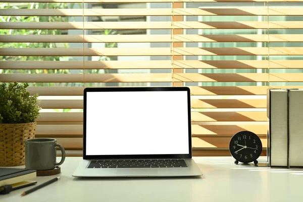 Mockup computer laptop with white empty display, alarm clock, books and potted plat on white table.