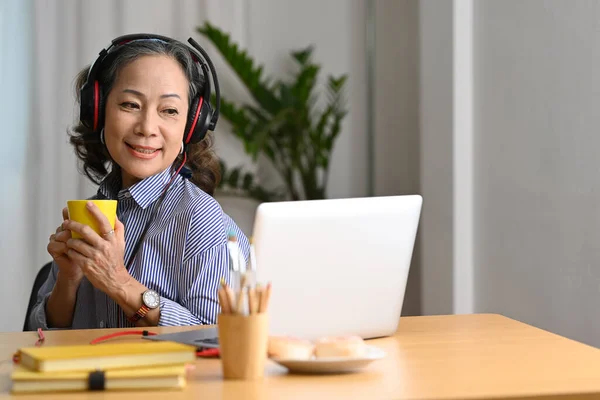 Smiling middle aged lady in headphone drinking coffee and watching online webinar, working at home with a laptop computer.