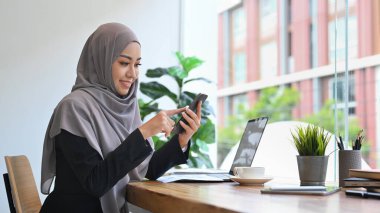 Ofiste bilgisayarla çalışırken cep telefonundan mesaj atan gülümseyen Müslüman kadın..