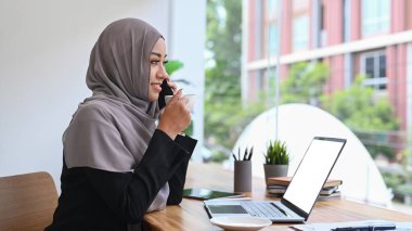  Başörtüsü takan Müslüman bir kadın telefonda konuşuyor ve modern ofis odasında sıcak kahve içiyor..