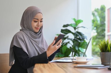 Başörtüsü takan Müslüman iş kadını dizüstü bilgisayarın önünde oturuyor ve cep telefonu kullanıyor..