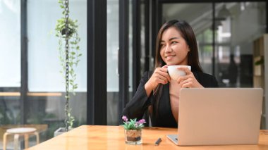 Tatmin olmuş genç girişimci iş yerinde oturup pencereden dışarı bakıyor ve gelecekteki başarılı projelerin hayalini kuruyor..