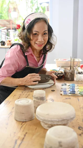 Happy middle aged woman decorating pottery before baking in workshop. Activity, handicraft, hobbies concept.