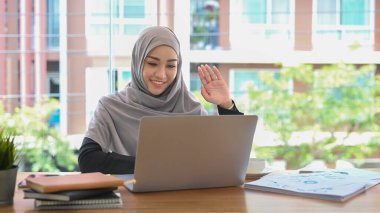 Türbanlı genç Müslüman iş kadını, modern işyerinde otururken bilgisayarında online video görüşmesi yapıyor..