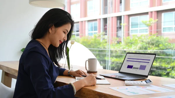 Side view of asian female entrepreneur sitting near large window in modern office and working with laptop computer.
