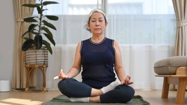 Nilüfer yogası pozisyonunda oturmuş oturma odasında meditasyon yapan orta yaşlı bir kadının tam boy portresi..