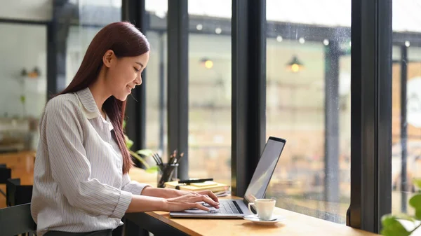 Smiling millennial female employee work on computer laptop in modern coffee shop.