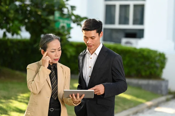Focused mature business leader and young businessman discussing online information, standing outside modern office building.