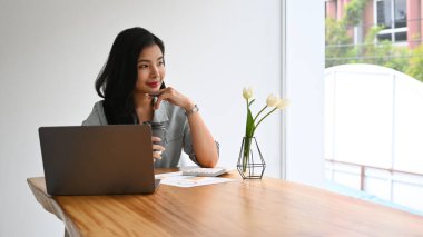 Düşünceli iş kadını dizüstü bilgisayarın önünde oturup pencereden dışarı bakıyor ve yeni başarılı projelerin hayalini kuruyor..