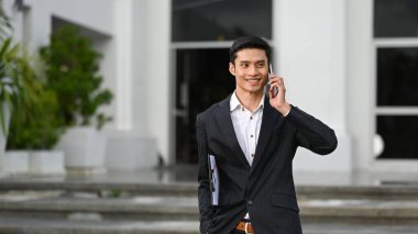 Resmi takım elbiseli yakışıklı bir işadamı cep telefonuyla konuşuyor ve ofis geçmişiyle dışarıda duruyor..