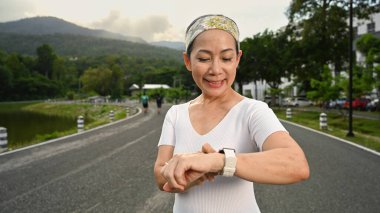 Smiling healthy middle aged woman checking heart rate data on smartwatch during jogging in the morning.