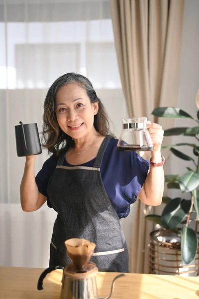 快乐的退休女人拿着黑色钢壶 对着相机笑着 在舒适的家里度过了一个清早 — 图库照片